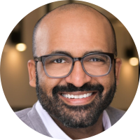 Brown skinned male with dark eyes behind black, square-rimmed glasses, minimal hair, but dark neatly trimmed beard.